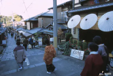 A narrow, sloped street in Japan lined with traditional wooden buildings. People wearing casual and traditional clothing walk past a café with white paper umbrellas displayed outside.