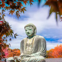 Kamakura Day Trip A large bronze Buddha statue sits serenely against a vibrant backdrop of autumn foliage with red and orange leaves. The sky is partly cloudy, providing a peaceful, tranquil atmosphere.