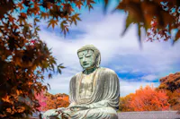A large bronze Buddha statue sits serenely against a vibrant backdrop of autumn foliage with red and orange leaves. The sky is partly cloudy, providing a peaceful, tranquil atmosphere.