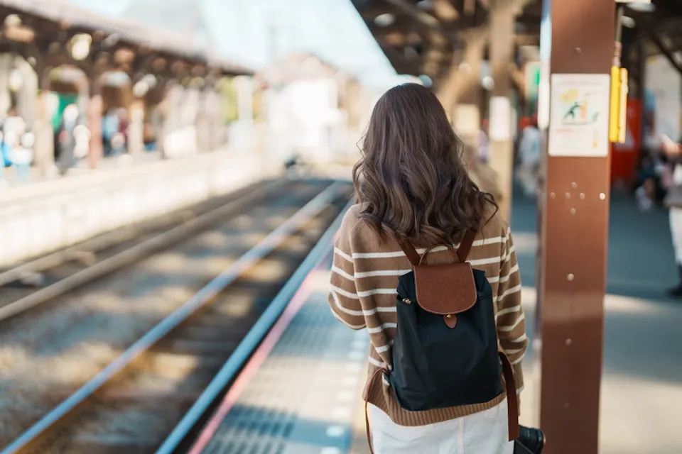 A person with wavy brown hair and a striped sweater stands on a train platform, facing away. They are wearing a black backpack and waiting beside the tracks. The platform is lit by daylight and appears to be in a busy railway station.