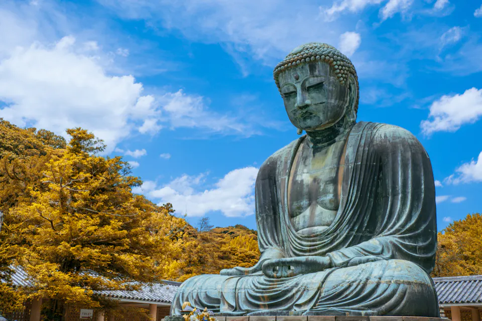 A large outdoor statue of Buddha sits serenely against a backdrop of leafy trees and a partly cloudy sky. The foliage is in shades of golden yellow, contrasting with the vivid blue of the sky. The statue’s expression is calm and contemplative.