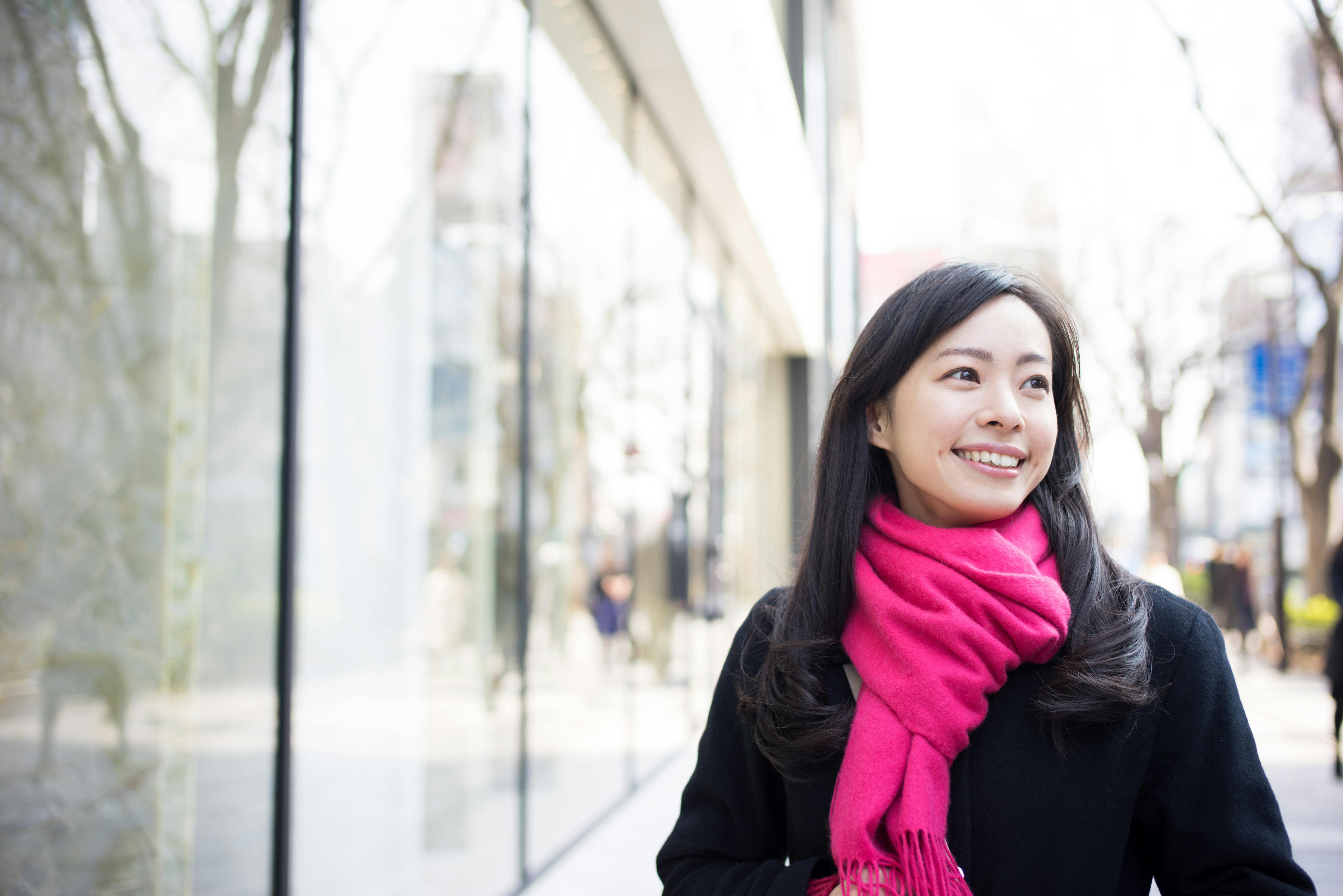 A woman with long dark hair, wearing a bright pink scarf and a black coat, smiles while standing outside next to large glass windows on a city street. Trees and blurred people are visible in the background.