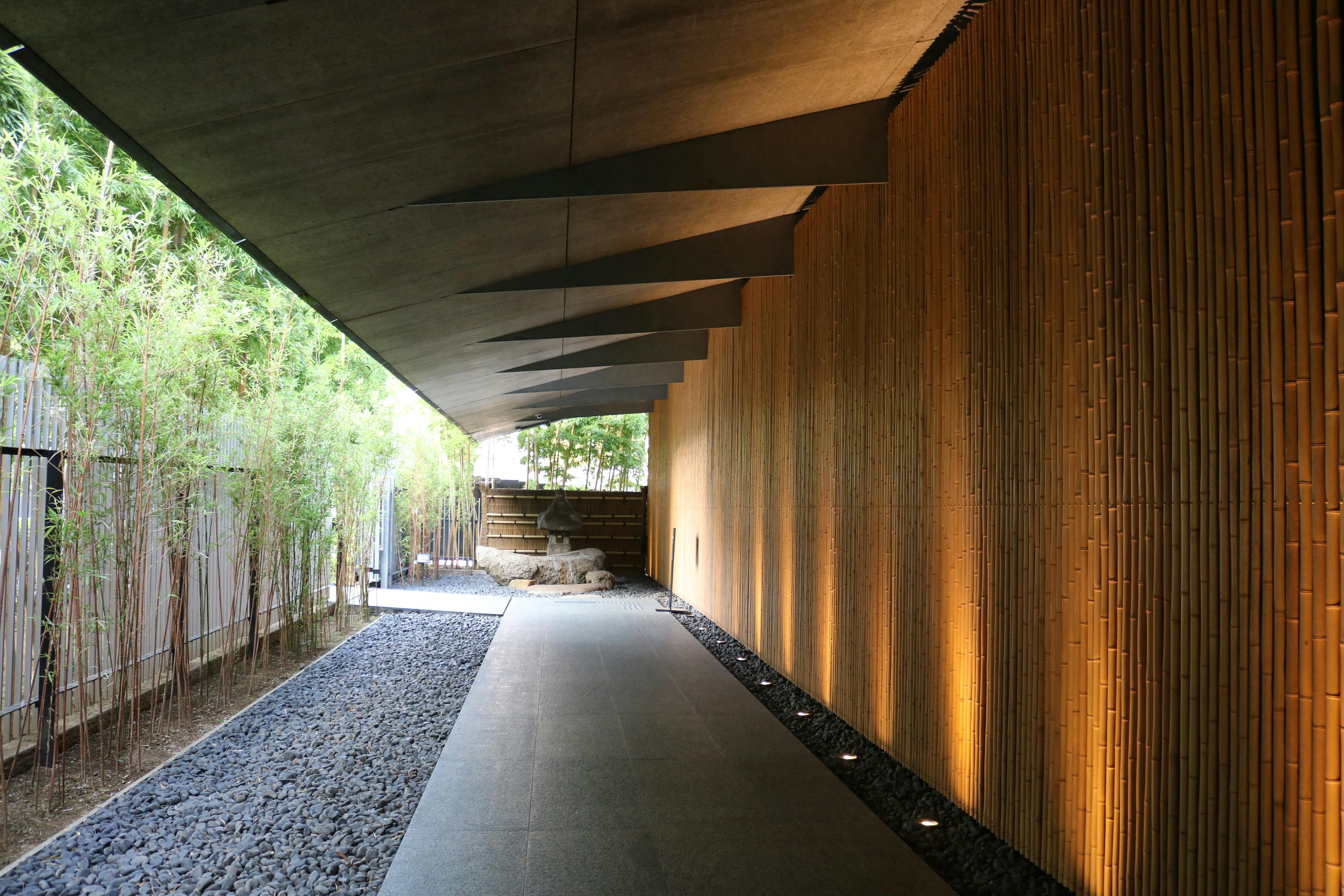 A modern covered walkway with a bamboo wall on the right, a row of spotlights lighting it, a gravel path, and bamboo plants on the left, leading to an open, sunlit area with rocks.