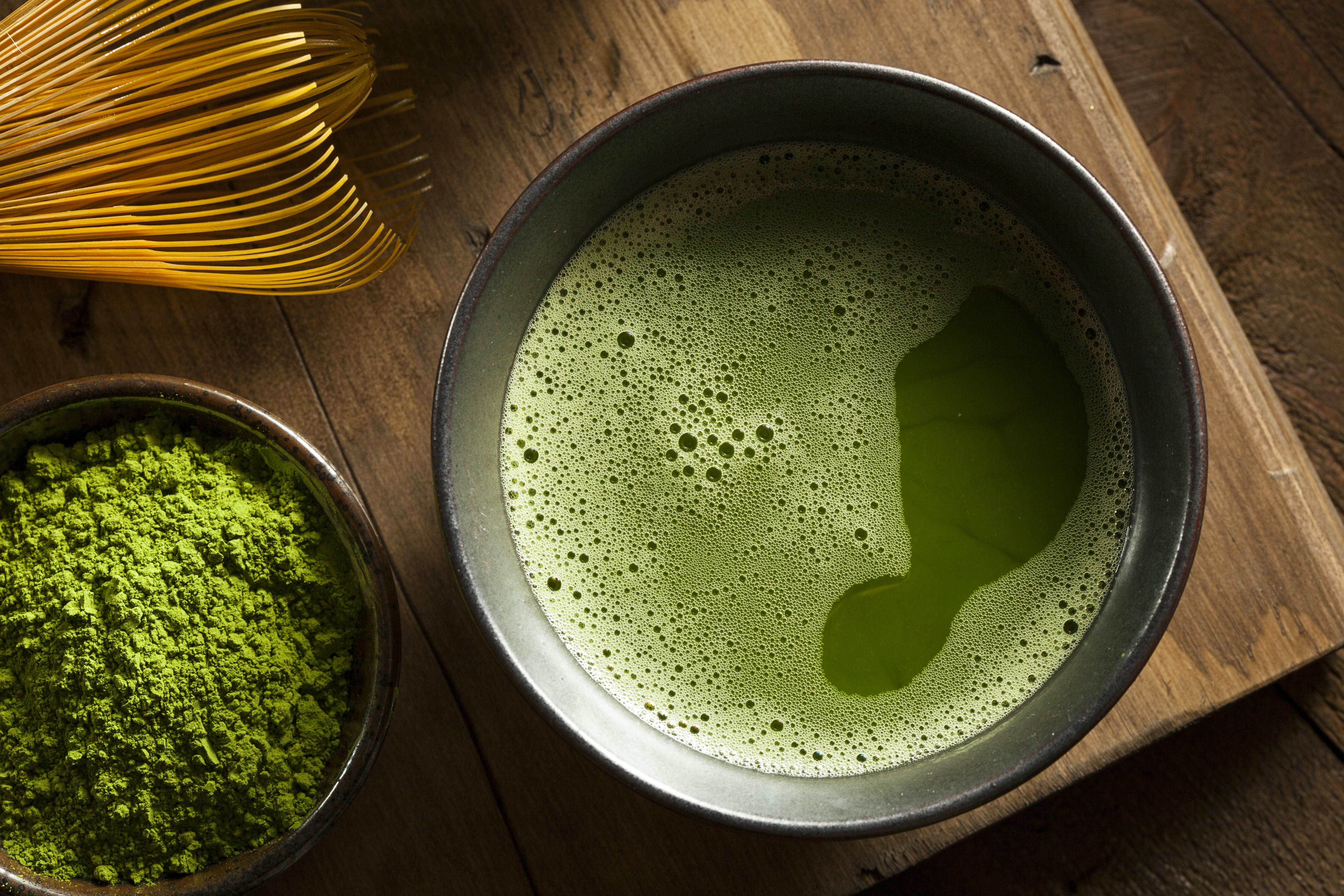 A bowl of frothy green matcha tea sits next to a small dish of vibrant matcha powder and a bamboo whisk on a wooden surface.