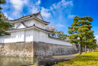 A traditional Japanese castle building with white walls and a tiled roof sits beside a stone wall and moat, surrounded by trimmed pine trees under a bright blue sky with scattered clouds.