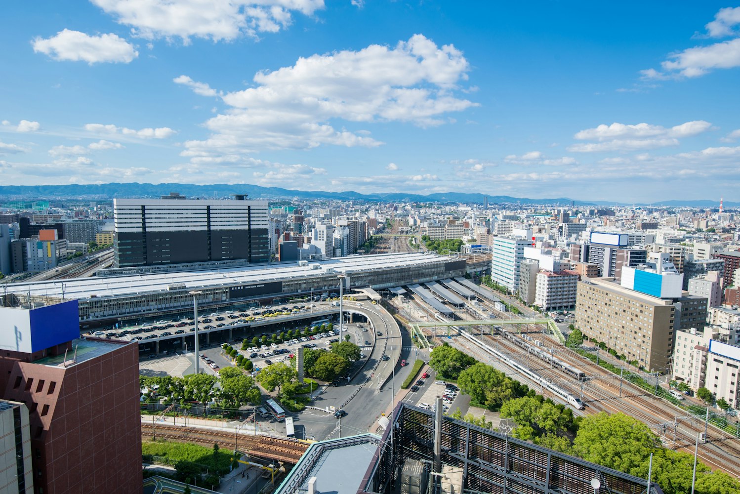 Aerial view of a bustling cityscape featuring a large train station with multiple tracks, surrounded by modern buildings, lush green spaces, and roads. The sky is clear and blue with scattered fluffy clouds.