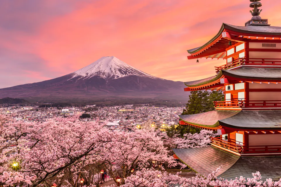 A beautiful view of Mount Fuji with a traditional Japanese pagoda and cherry blossoms in the foreground. The sky is painted with vibrant pink and orange hues during sunset.