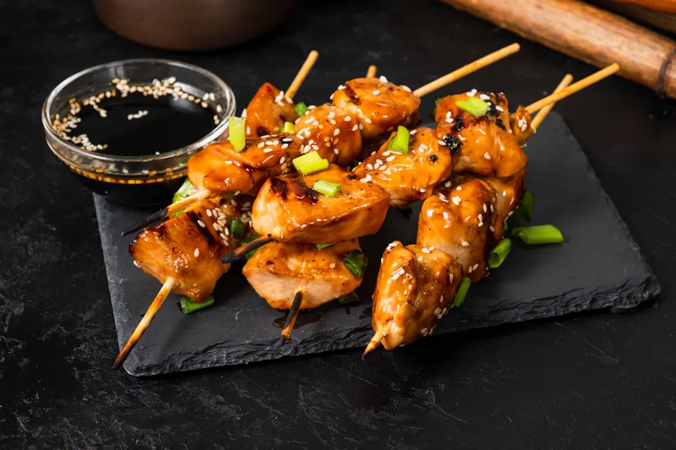 Skewered grilled chicken pieces glazed with a shiny sauce, garnished with sesame seeds and chopped green onions, served on a black slate plate with a small bowl of soy sauce on the side.