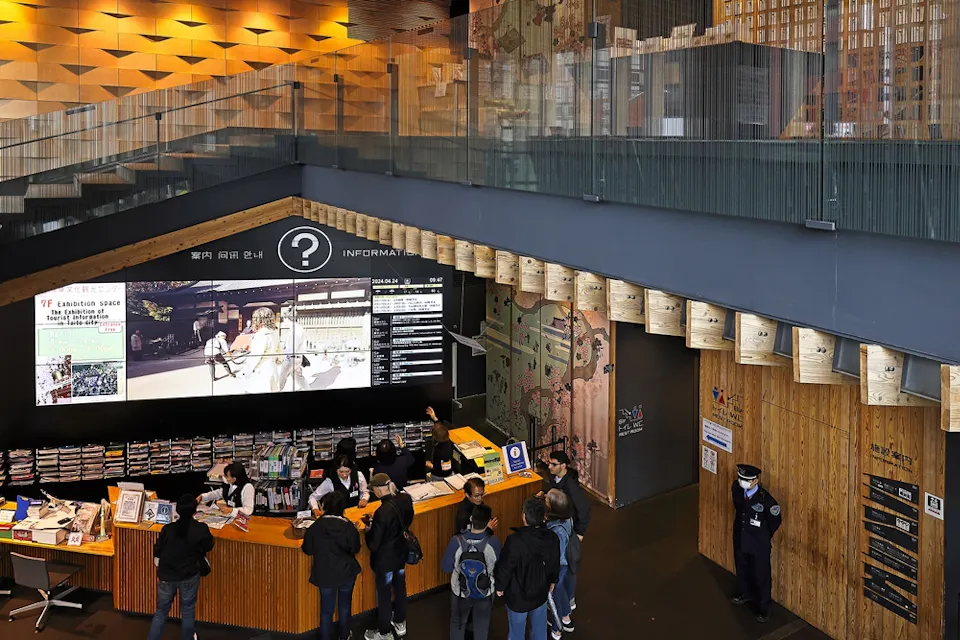 A modern information center with a high ceiling and wooden accents. People gather at a circular desk for assistance. An electronic display shows various information, and a staff member stands nearby.