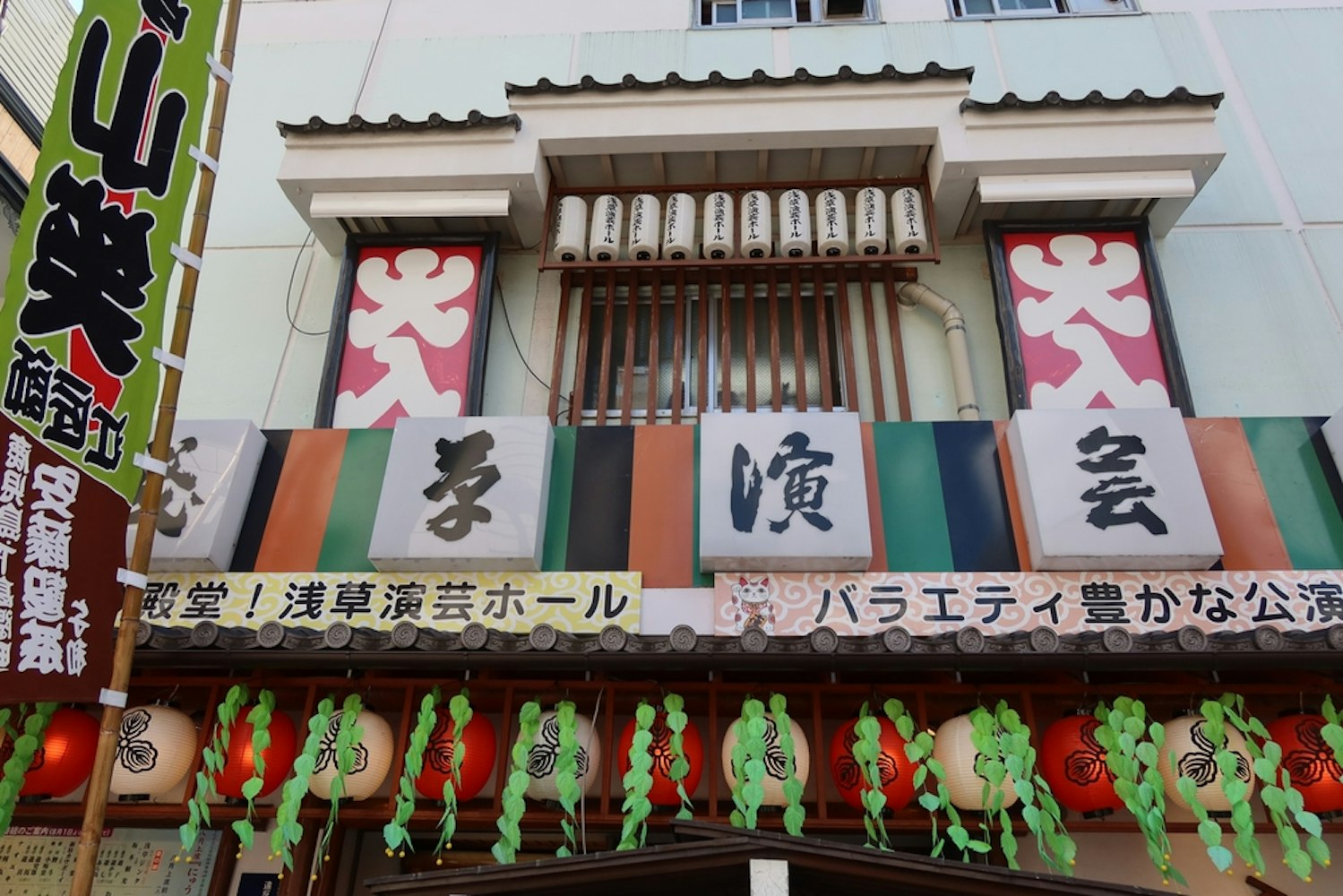 A colorful building facade in Japan displays traditional lanterns and signs with Japanese characters. Decorative green foliage hangs below, adding a festive look to the scene.