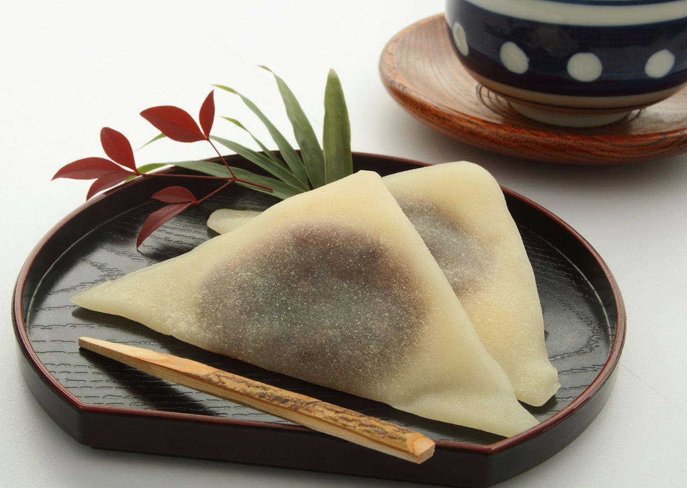 Two pieces of Yatsuhashi, Japanese sweets with a triangle shape and a filling, are placed on a black tray. A decorative branch and a small wooden utensil are beside them. In the background, a patterned bowl on a wooden base is visible.