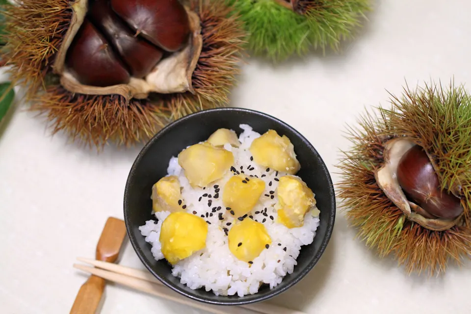 A bowl of rice topped with yellow chestnuts and sprinkled with black sesame seeds is in the center, surrounded by fresh chestnuts in their spiky shells. Wooden chopsticks rest nearby.