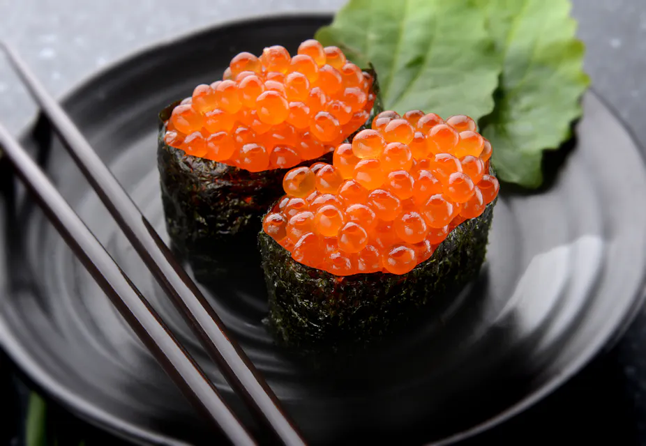 Two pieces of ikura sushi, topped with vibrant orange salmon roe, sit on a black plate garnished with green leaves. A pair of wooden chopsticks rests beside the dish.