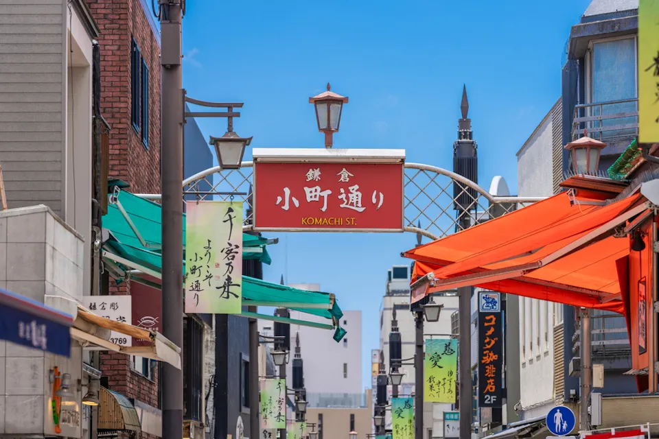 A vibrant street scene in Japan features a red archway sign reading "Komachi St." in both Japanese and English. Colorful banners and awnings line the street, and people walk below a clear blue sky.
