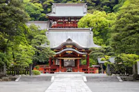 A traditional Japanese shrine surrounded by lush green trees. The building features red wooden structures and ornate roof designs, set against a backdrop of dense greenery. Stone paths lead to the entrance.