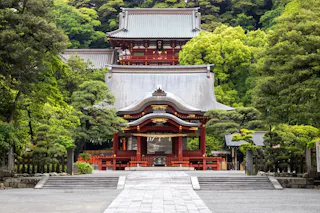 A traditional Japanese shrine surrounded by lush green trees. The building features red wooden structures and ornate roof designs, set against a backdrop of dense greenery. Stone paths lead to the entrance.
