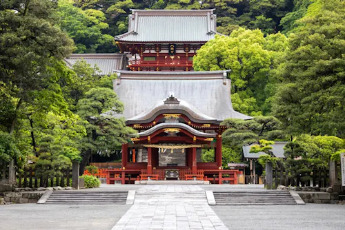 A traditional Japanese shrine surrounded by lush green trees. The building features red wooden structures and ornate roof designs, set against a backdrop of dense greenery. Stone paths lead to the entrance.
