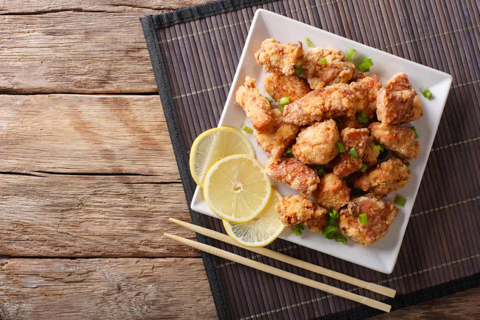 Spicy chicken karaage with lemon and green onion A square white plate on a bamboo mat holds crispy fried chicken pieces garnished with chopped green onions. Two lemon slices are placed on the side. Wooden chopsticks rest beside the plate on a rustic wooden surface.