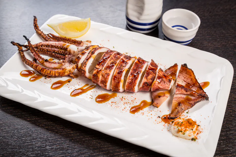 Japanese grilled squid ikayaki izakaya Grilled squid sliced on a rectangular white plate, garnished with a lemon wedge, sauce drizzle, and a dollop of seasoned mayonnaise. Two striped condiment containers are visible in the background on a dark wooden surface.