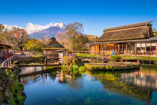 Traditional Japanese buildings with thatched roofs beside a serene pond, lush greenery, a small wooden bridge, and Mount Fuji with a snow-capped peak in the background under a clear blue sky.