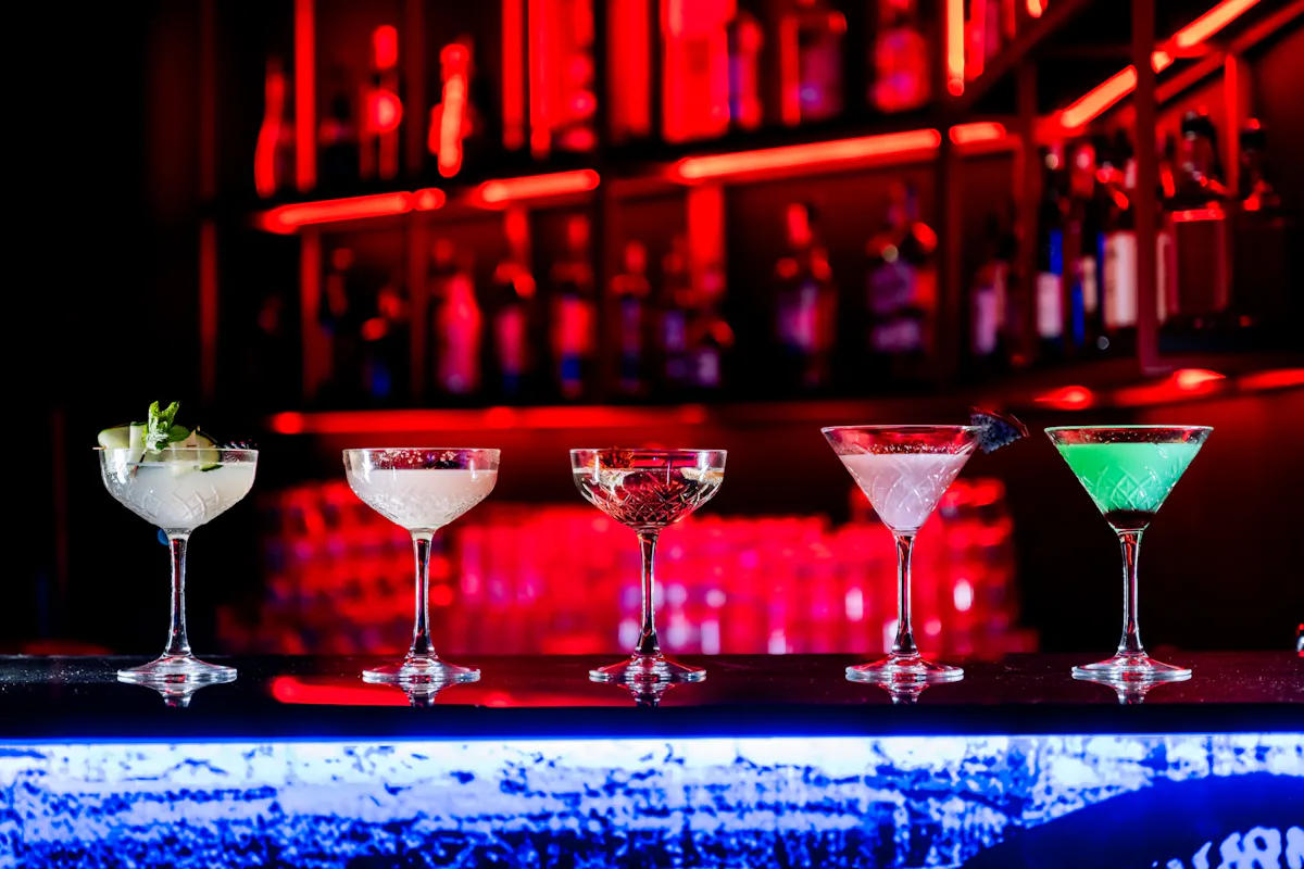 A row of five elegant cocktails in martini glasses is displayed on a dimly lit bar counter. Each drink varies in color and garnish, set against a backdrop of shelves with illuminated red lights and bottles.
