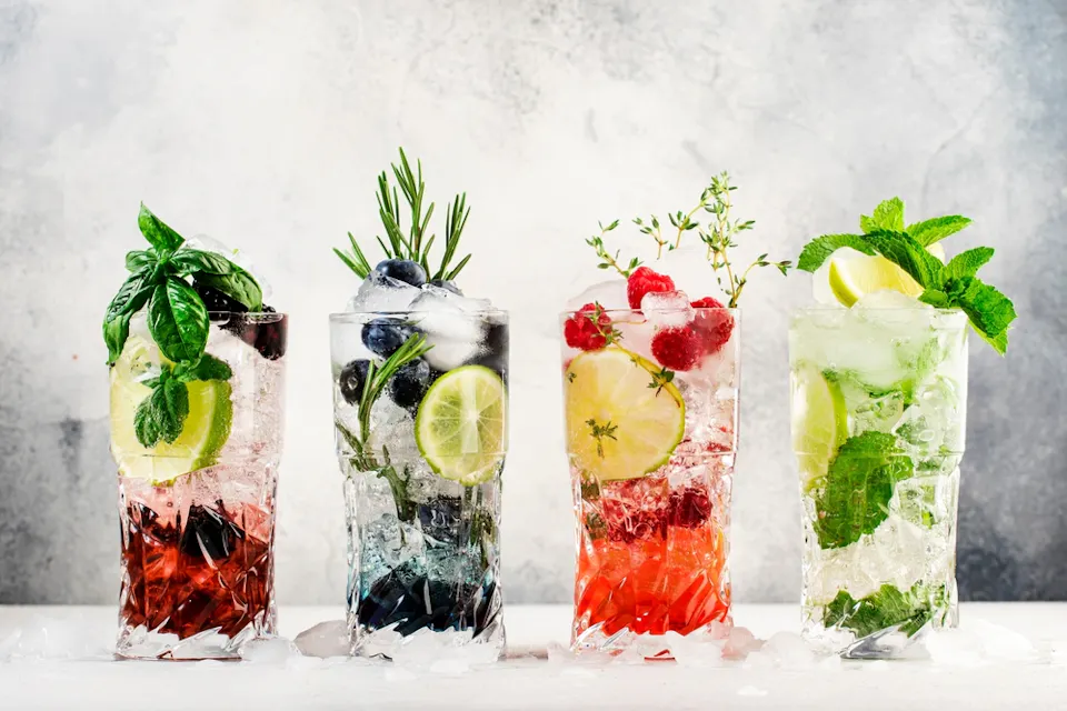 Four refreshing cocktails in tall glasses with ice, each garnished differently. From left to right: mint and lime, rosemary and blueberries, lemon and raspberries, mint and lime slices. The background is a light marbled surface.