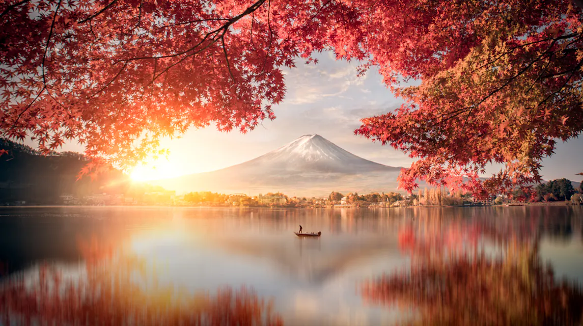 Mount Fuji in Autumn A calm lake reflects a boat with two people and the distant snow-capped Mount Fuji. Vibrant red and orange autumn leaves frame the scene, with the sun casting a warm glow over the tranquil landscape.