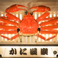 Giant Crab in Osaka's Restaurant A large, vibrant red crab sculpture is displayed on the exterior of a building, illuminated against a striped backdrop. Japanese characters are visible beneath the sculpture, likely indicating the name of a shop or restaurant.