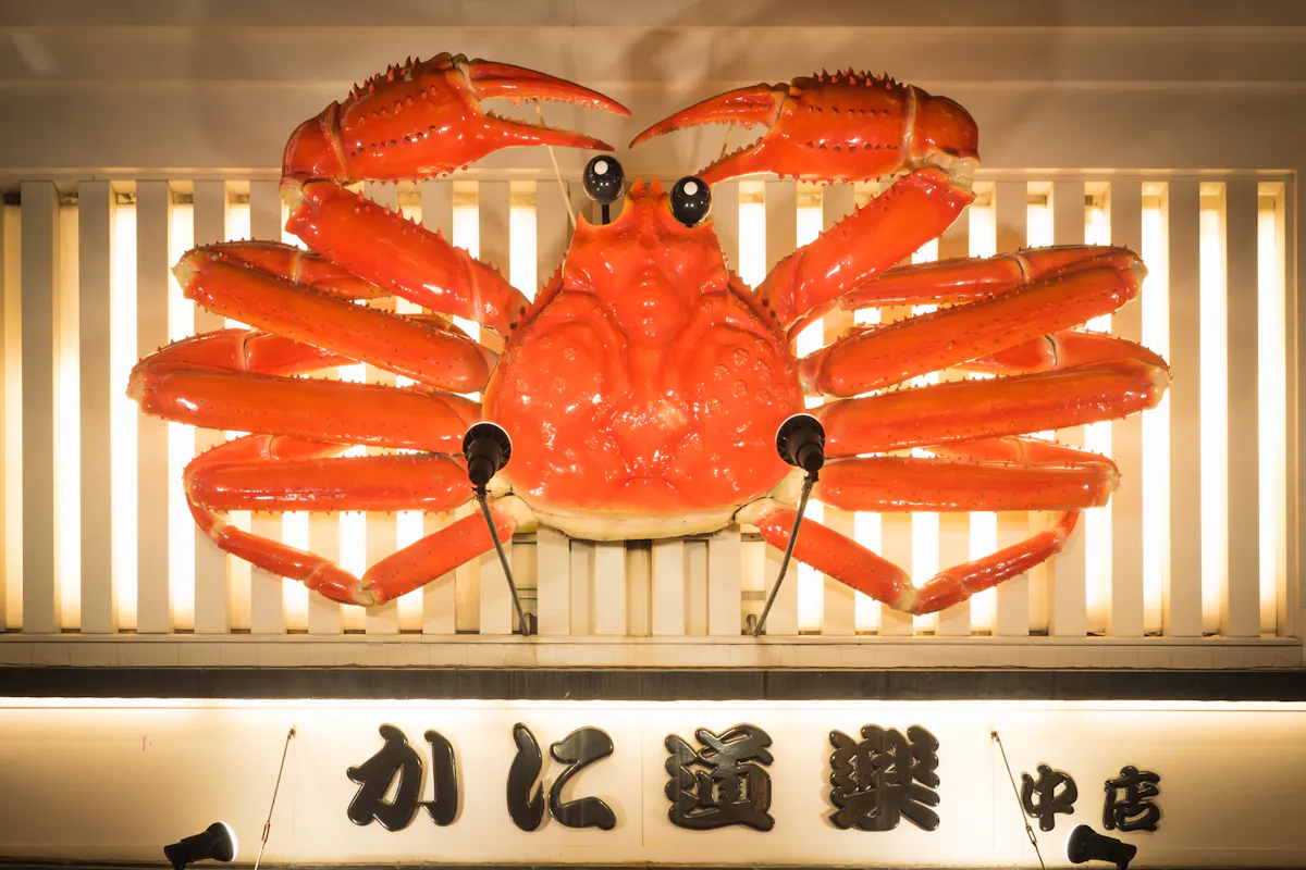 Giant Crab in Osaka's Restaurant A large, vibrant red crab sculpture is displayed on the exterior of a building, illuminated against a striped backdrop. Japanese characters are visible beneath the sculpture, likely indicating the name of a shop or restaurant.