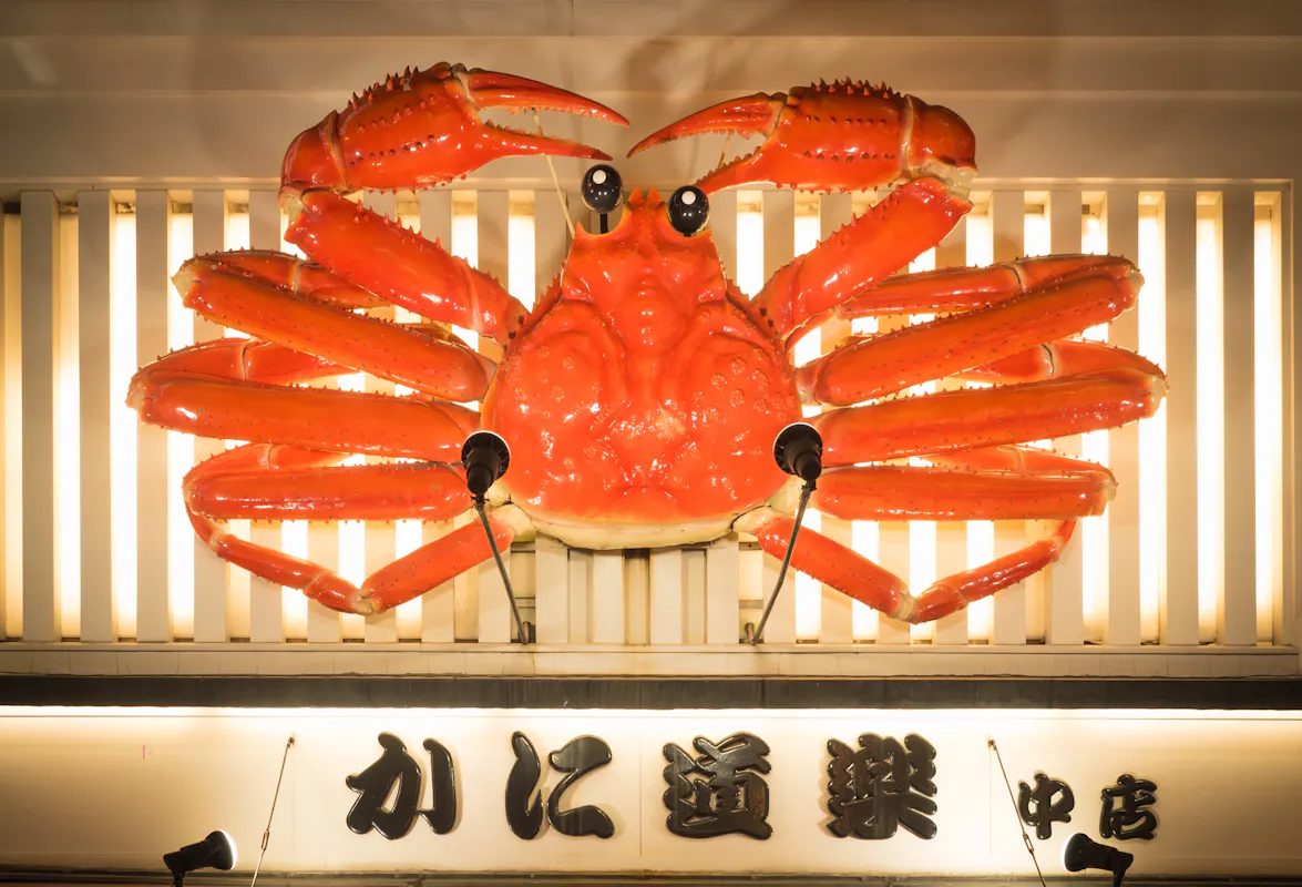 A large, vibrant red crab sculpture is displayed on the exterior of a building, illuminated against a striped backdrop. Japanese characters are visible beneath the sculpture, likely indicating the name of a shop or restaurant.
