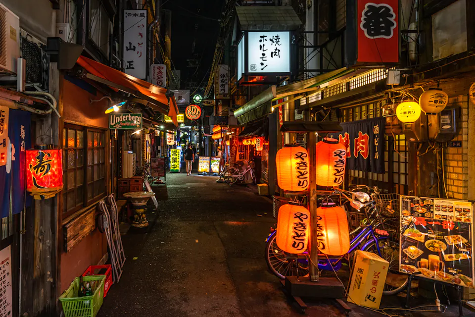 Nighttime alley in Japan, illuminated by vibrant red and orange lanterns and neon signs. The narrow street is lined with small, cozy restaurants and bicycles are parked along the walkway, creating a warm, inviting atmosphere.