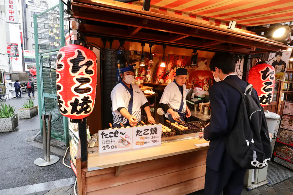 A street food stall with lanterns selling takoyaki is staffed by two people wearing uniforms and hats. A customer stands in front, looking at the offerings and prices displayed on signs. Urban setting with buildings visible in the background.