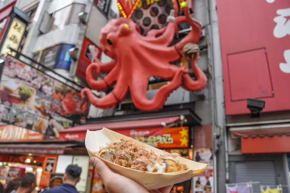 A hand holds a boat of takoyaki in the foreground, topped with bonito flakes and sauce. In the background, a large octopus sculpture is mounted above a bustling street food stall, creating a vibrant and busy street scene.
