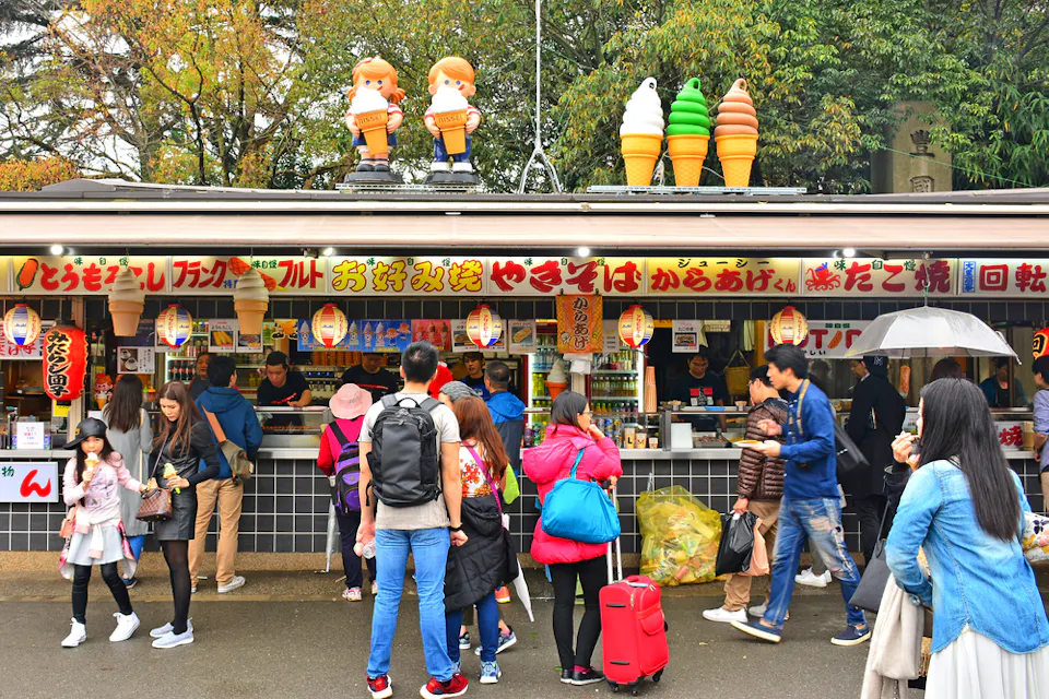People standing in line at a colorful food stall decorated with large ice cream cones and cartoon figures. The stall has Japanese signs and offers snacks and drinks. Customers are engaging with vendors under a cloudy sky.