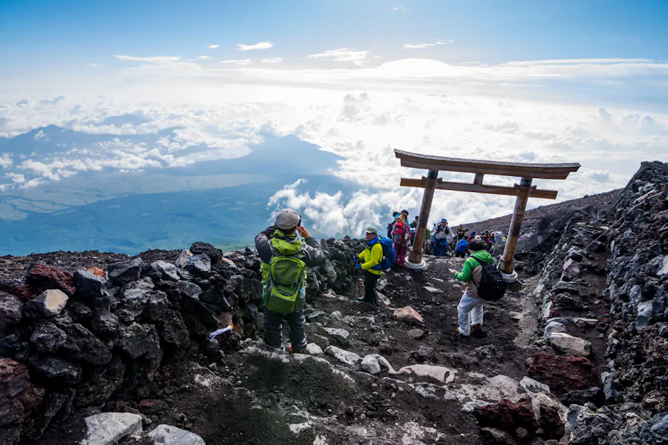 Hiking Mt Fuji A group of hikers descends a rocky path on a mountainside under a clear sky. A traditional wooden torii gate stands on the trail. The background showcases clouds and expansive landscape views.