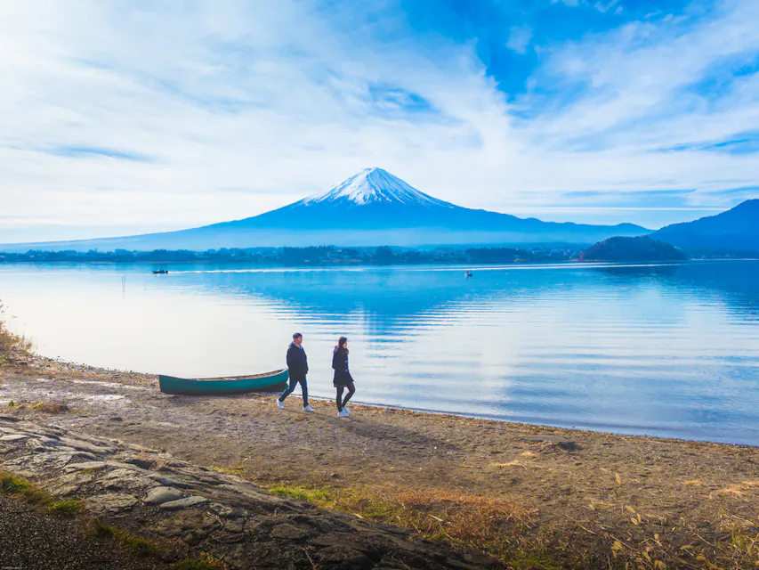 Mt. Fuji Two people walk along a lakeshore under a clear blue sky, with a small boat nearby. In the background, Mount Fuji rises majestically, reflecting in the calm water.