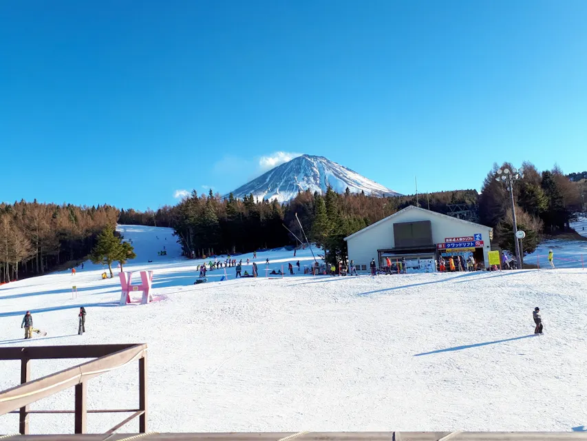 A ski resort with a snow-covered slope, people skiing, and a building nearby. In the background, there's a snow-capped mountain under a clear blue sky.