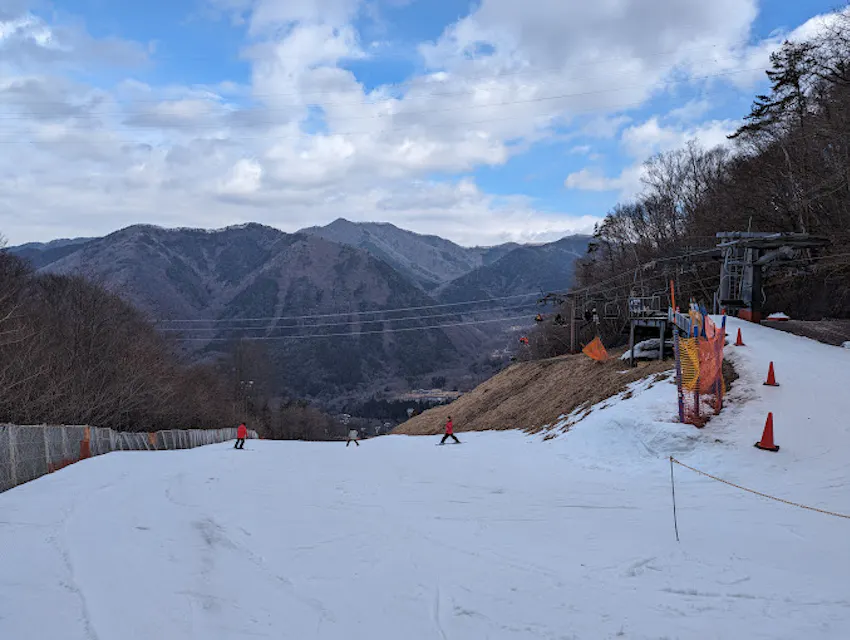 A ski slope with skiers descending, surrounded by snow on a clear day. Mountains and a partly cloudy sky are in the background, with a ski lift to the right. Orange cones and fencing line part of the snowy path.