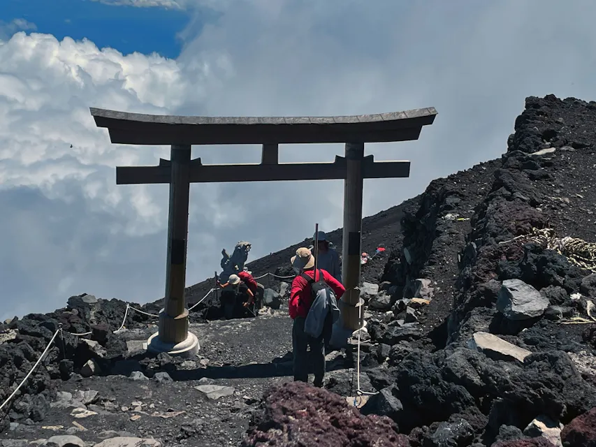 Two hikers walk through a wooden torii gate on a rocky mountain trail. The landscape is rugged, with a dramatic cloud-filled sky in the background, suggesting high altitude and a remote location.