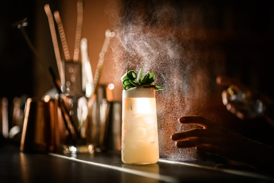 A cocktail in a tall glass with ice, garnished with green leaves, on a bar counter. A hand is seen misting the drink, with a soft glow and blurred background showcasing bar utensils.