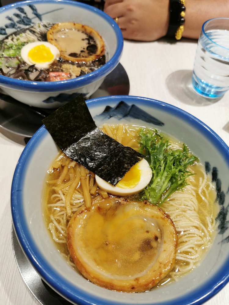 Two bowls of ramen are on a table. The front bowl contains noodles, a slice of chashu, half a boiled egg, bamboo shoots, seaweed, and greens in a clear broth. The bowl in the background has darker broth and similar toppings. A glass of water is nearby.