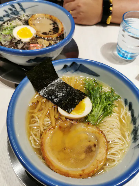 Yuzu Shio Ramen Two bowls of ramen are on a table. The front bowl contains noodles, a slice of chashu, half a boiled egg, bamboo shoots, seaweed, and greens in a clear broth. The bowl in the background has darker broth and similar toppings. A glass of water is nearby.