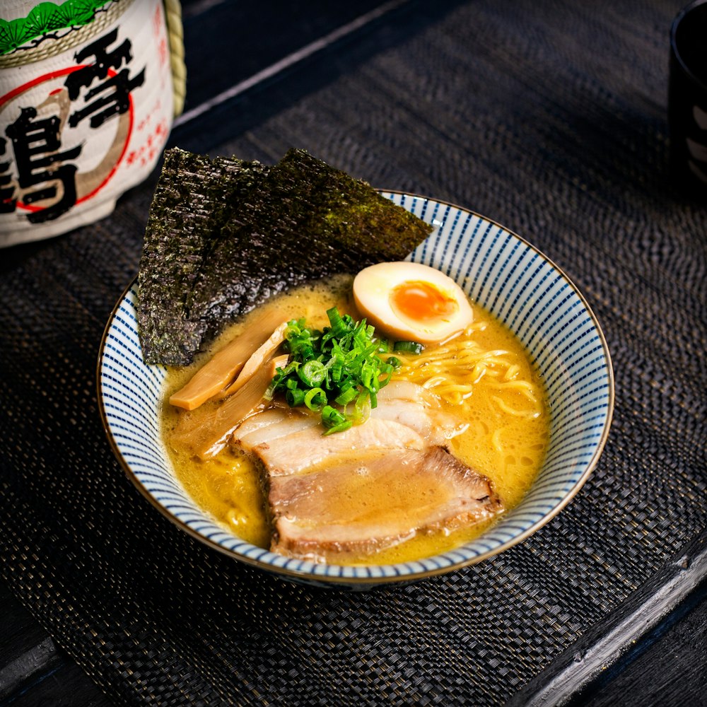 A bowl of ramen with rich broth, garnished with slices of pork, a halved soft-boiled egg, green onions, bamboo shoots, and nori sheets. The dish is served in a blue-striped bowl on a dark woven mat with Japanese decor in the background.