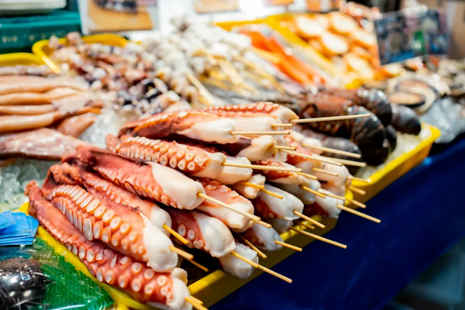 Skewered octopus tentacles on display at a seafood market, surrounded by a variety of fresh seafood items on ice in a yellow tray.