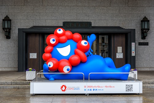 A vibrant blue mascot with red circular features and googly eyes lounges on a display platform. The backdrop is a stone building entrance with an EXPO 2025 Osaka, Kansai, Japan sign, featuring event dates and location details.