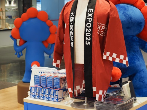 A store display features a red and black jacket with "EXPO 2025" printed on it. Nearby, there are stacks of boxed items and promotional materials. In the background, colorful abstract figures add a playful atmosphere.