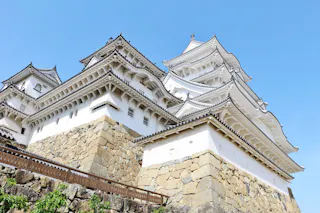 A white traditional Japanese castle with tiered roofs and ornate eaves stands atop a stone foundation against a clear blue sky.