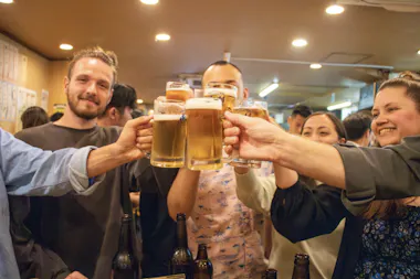A group of people smile and raise glasses of beer in a toast at a lively indoor gathering, with bottles visible on the table in front of them.