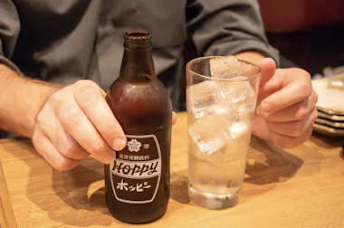 A person holds a dark glass bottle labeled "Hoppy" in one hand and a tall glass filled with ice in the other, sitting at a wooden table.