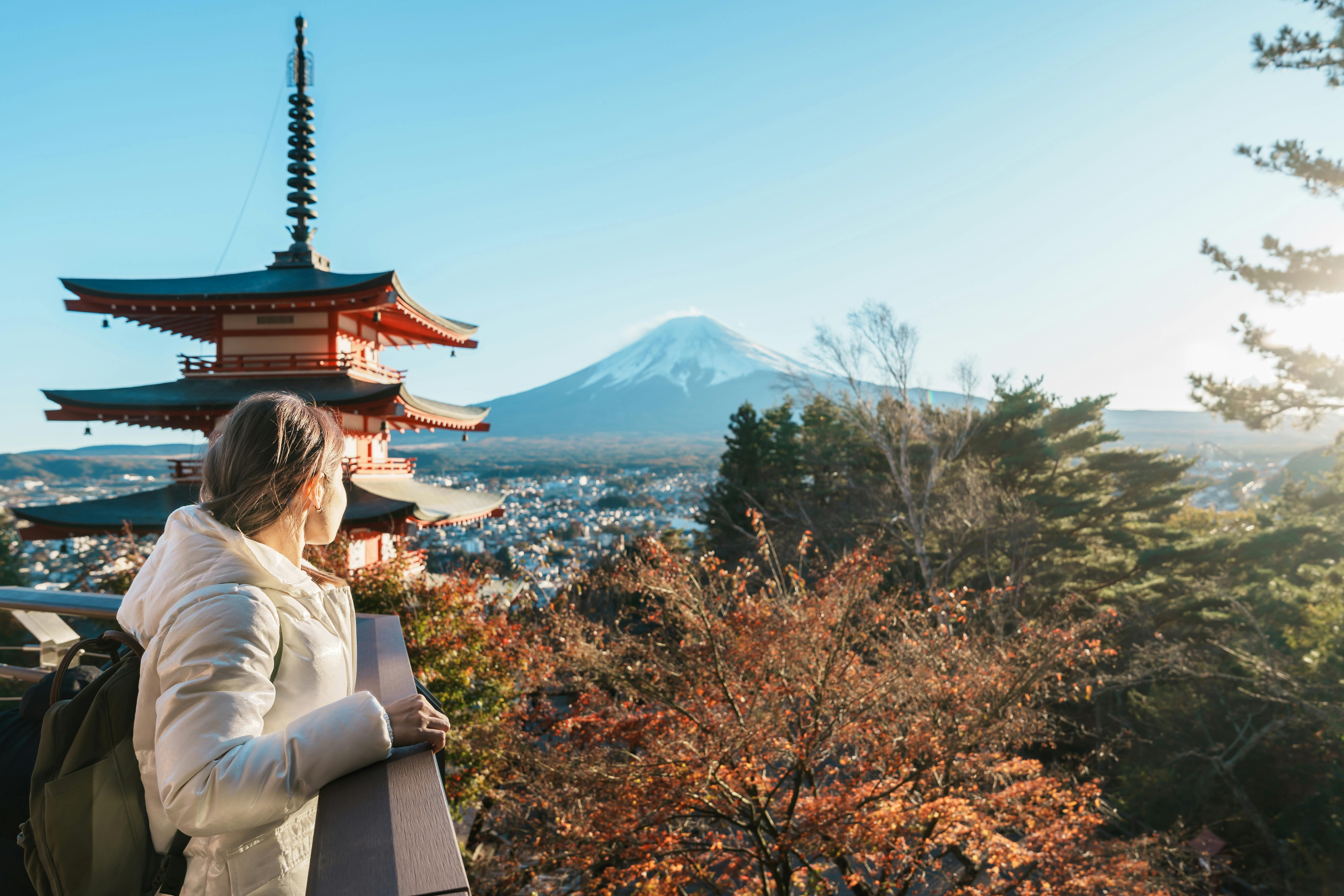 A person in a white coat stands on a balcony, overlooking autumn trees, a red pagoda, and Mount Fuji under a clear sky in Japan.